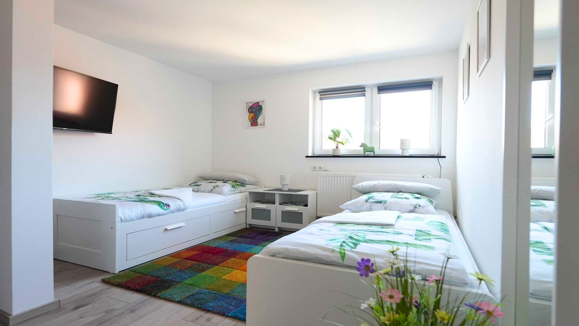 ferienwohnung-bad-koenigshofen-zweibettzimmer-hell-tv Zweibettzimmer einer Ferienwohnung in Bad Königshofen mit TV, Tageslicht und farbenfrohem Teppich.