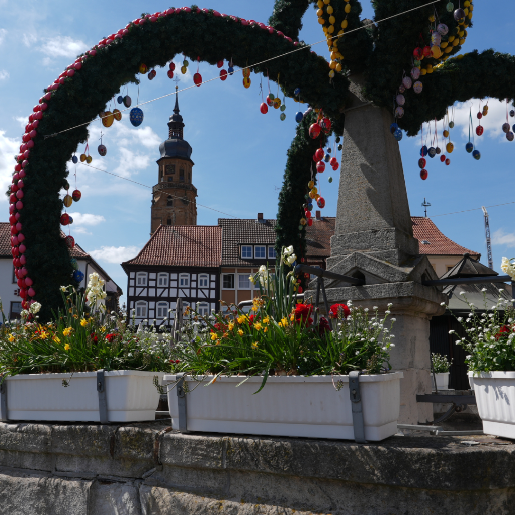 Osterbrunnen mit buntem Blumenschmuck und Blick auf den historischen Stadtturm in Bad Königshofen.