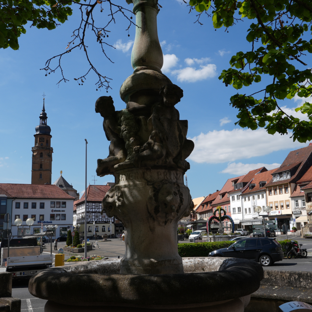 Historischer Brunnen im Schatten eines Baumes auf dem Marktplatz von Bad Königshofen, im Hintergrund Fachwerkhäuser und der markante Kirchturm unter blauem Himmel