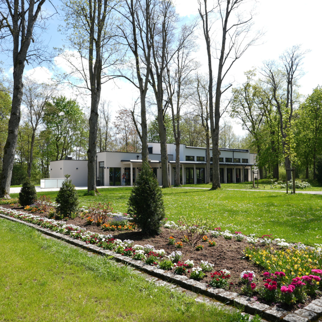 Blumenbeet mit bunten Frühlingsblumen vor der modernen Frankentherme in Bad Königshofen, umgeben von hohen Bäumen und gepflegtem Rasen im Kurpark