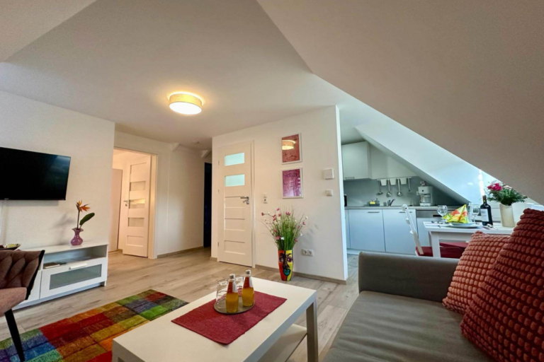 Heller Wohnraum mit Couch, Couchtisch und einladender Deko, angrenzend an eine moderne Küchenzeile unter der Dachschräge – farbenfrohe Akzente und offene Raumgestaltung.