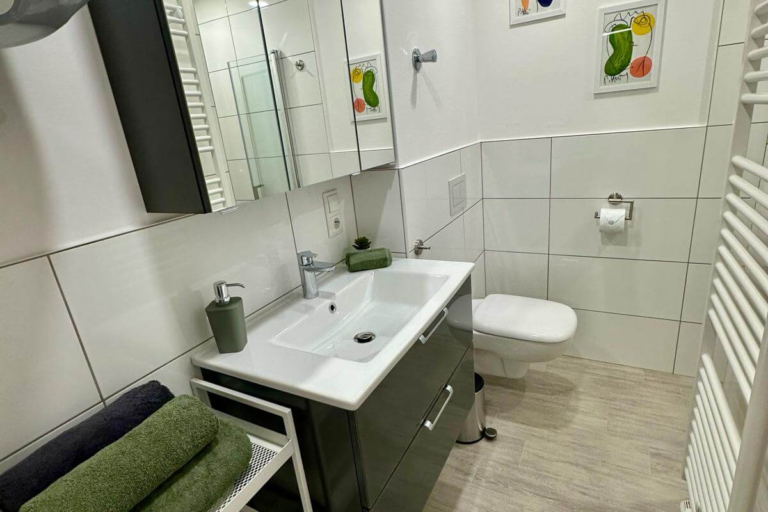 Badezimmer mit großzügigem Spiegel, Waschbecken mit dunklem Unterschrank, WC und stilvollen Akzenten in Grün – freundlich und modern eingerichtet.