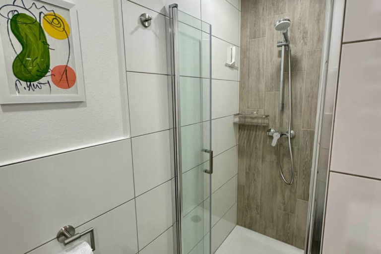 Helle, moderne Dusche mit Glaswand, weißen Fliesen und natürlicher Holzoptik – stilvoll und komfortabel ausgestattet.