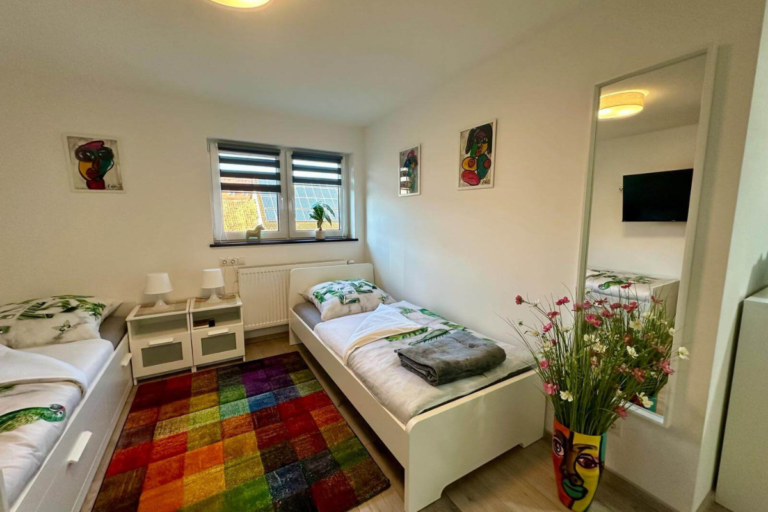 Gemütlich eingerichtetes Schlafzimmer mit zwei Einzelbetten, bunter Teppich und frischer Blumendekoration vor dem Fenster